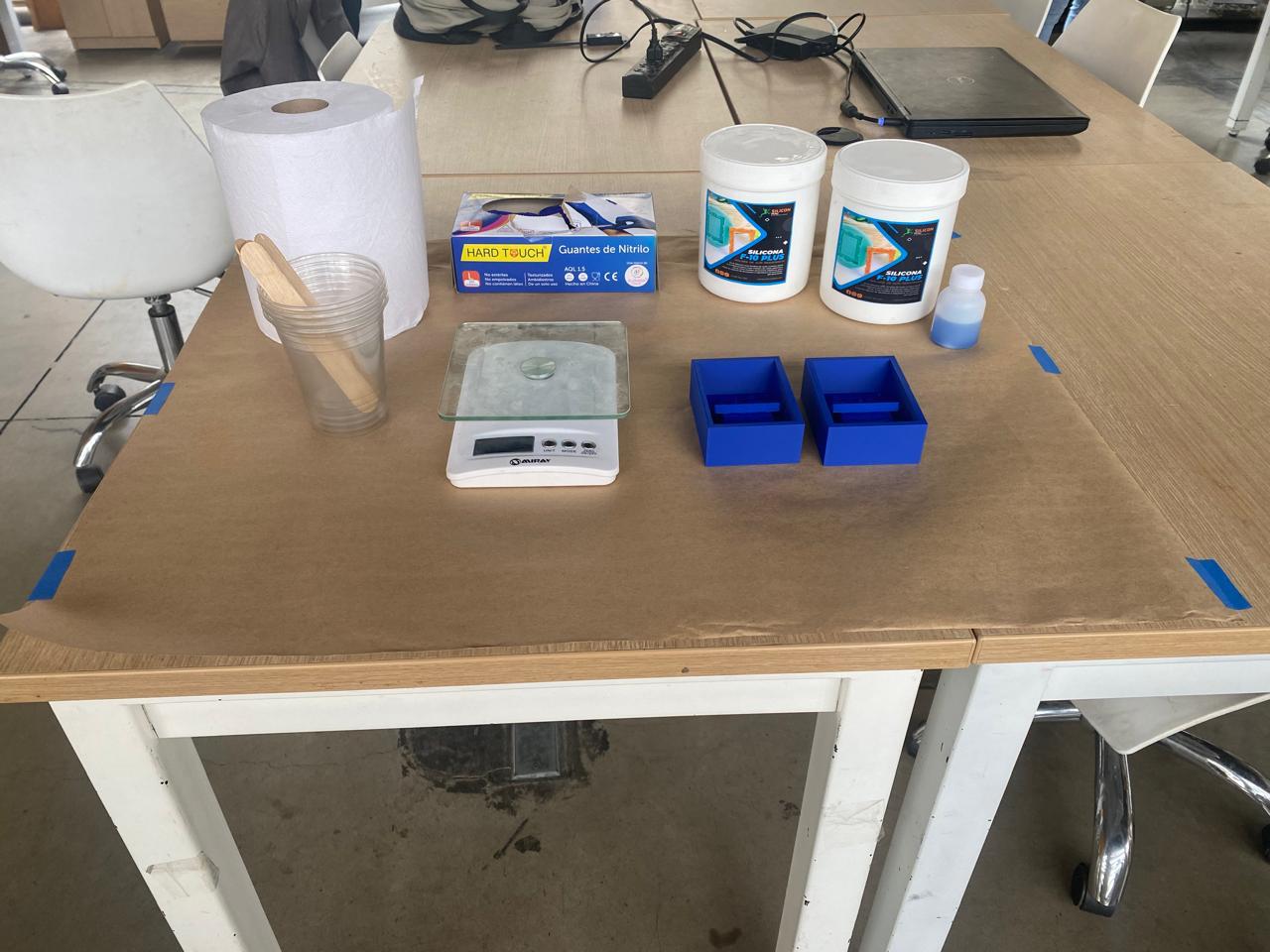 Workspace prepared — kraft paper, Silicona F-10 PLUS containers, scale, mixing cups, mold containers, and nitrile gloves laid out