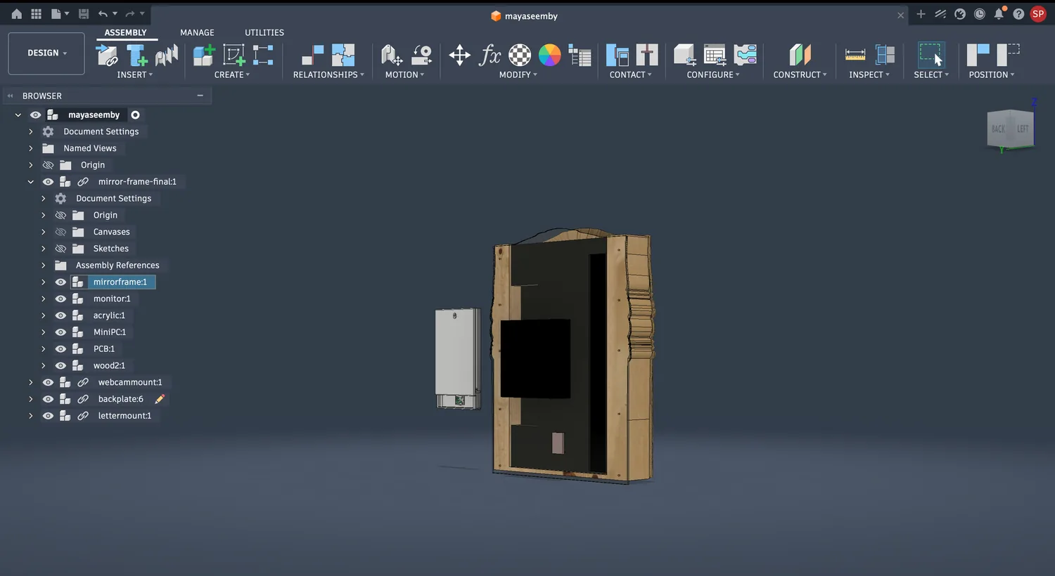 Fusion 360 back/side view of the assembly showing the screen cavity inside the wood frame, with the browser tree listing mirrorframe, monitor, acrylic, MiniPC, PCB, wood, webcammount, backplate and lettermount