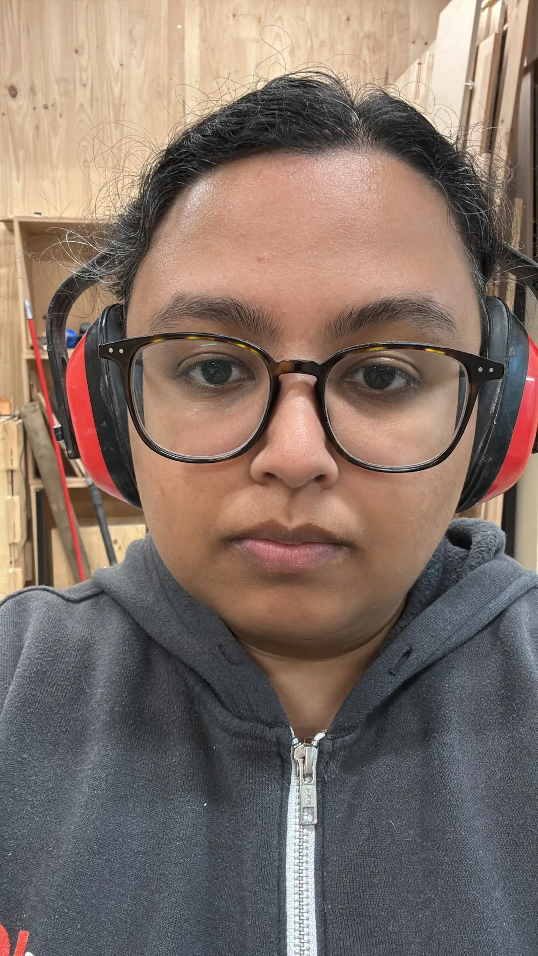 Self-portrait at the CNC in ear-protection muffs and safety glasses, timber stock on the racks behind