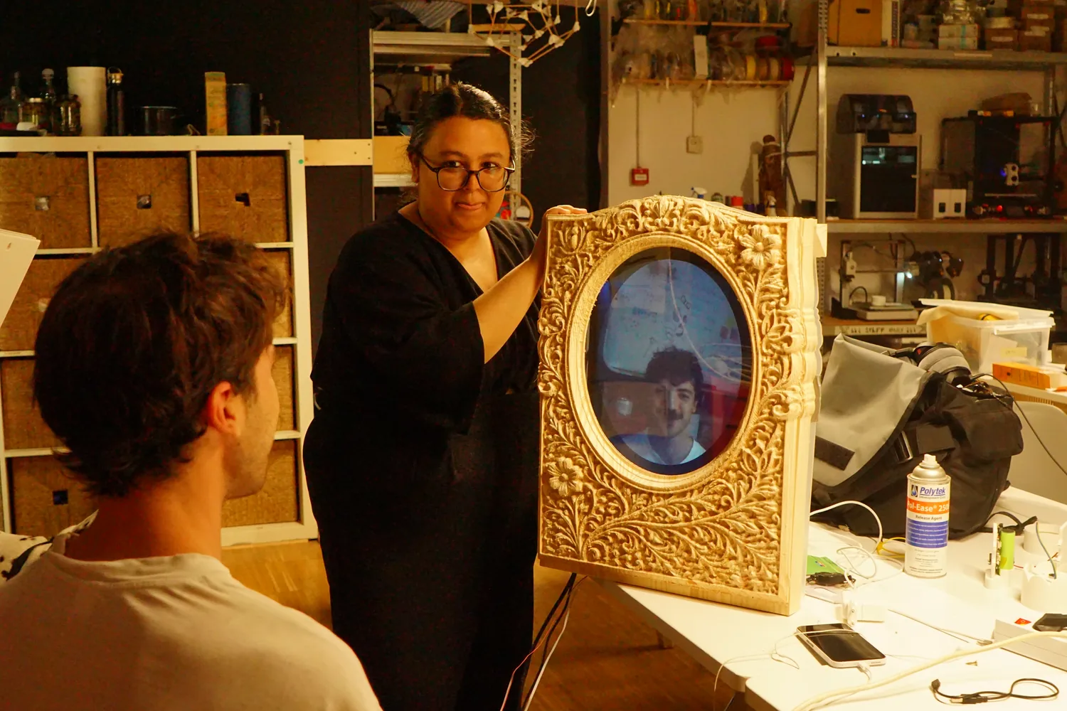 Maya — my grandmother's letter made tangible: a carved pine mirror on a white brick wall, a viewer's face in the oval, the lit acrylic letter beside it, two people steadying the frame