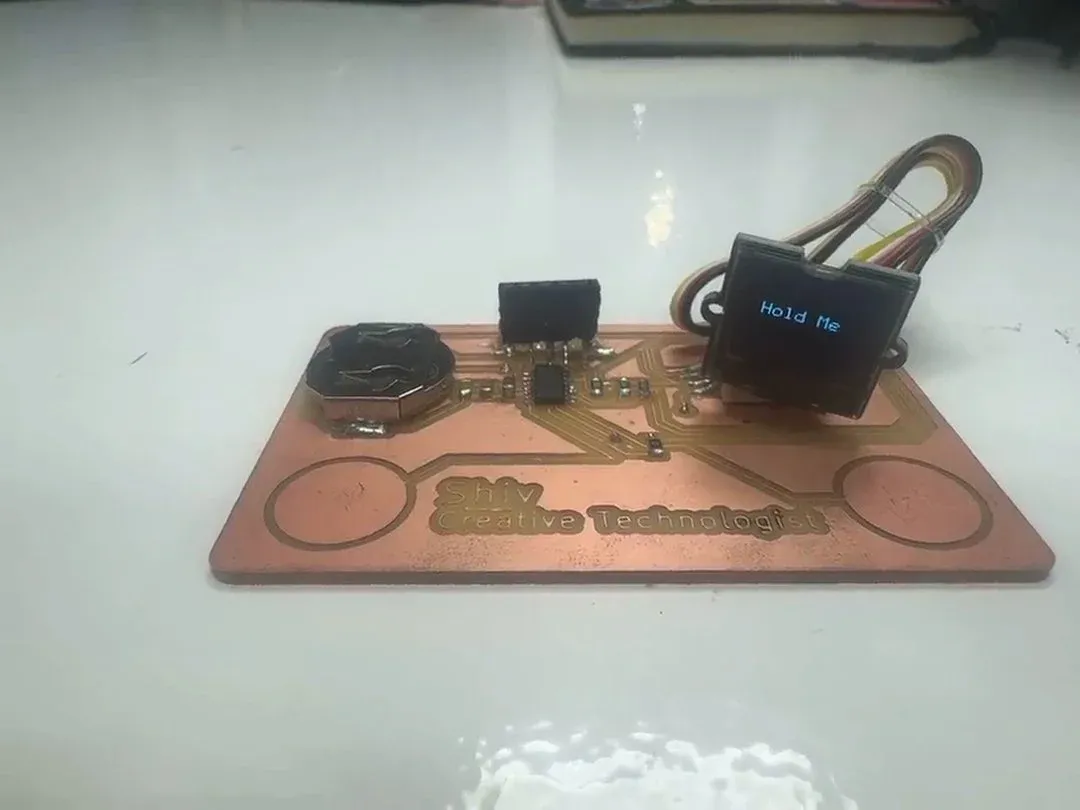 The PCB business card with the Grove SSD1306 OLED showing 'Hold Me' in blue, copper board engraved 'Shiv Creative Technologist', coin cell and ATSAMD11 visible