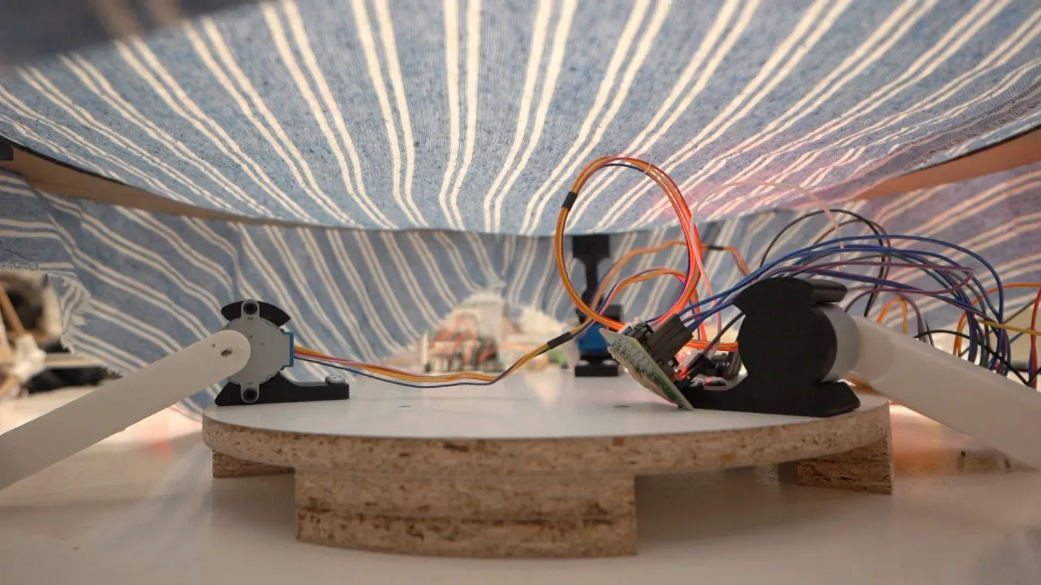 Low-angle view under the machine: three stepper-driven arms and the joystick beneath the blue striped cloth stretched across the ring