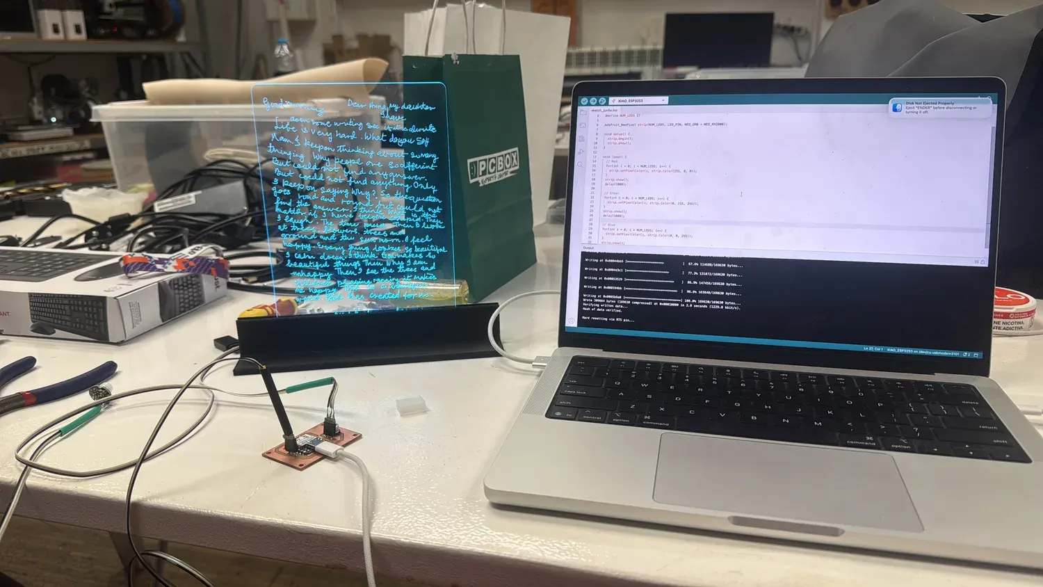 Edge-lit acrylic letter glowing blue on a stand, next to a laptop running the NeoPixel sketch flashing to the XIAO