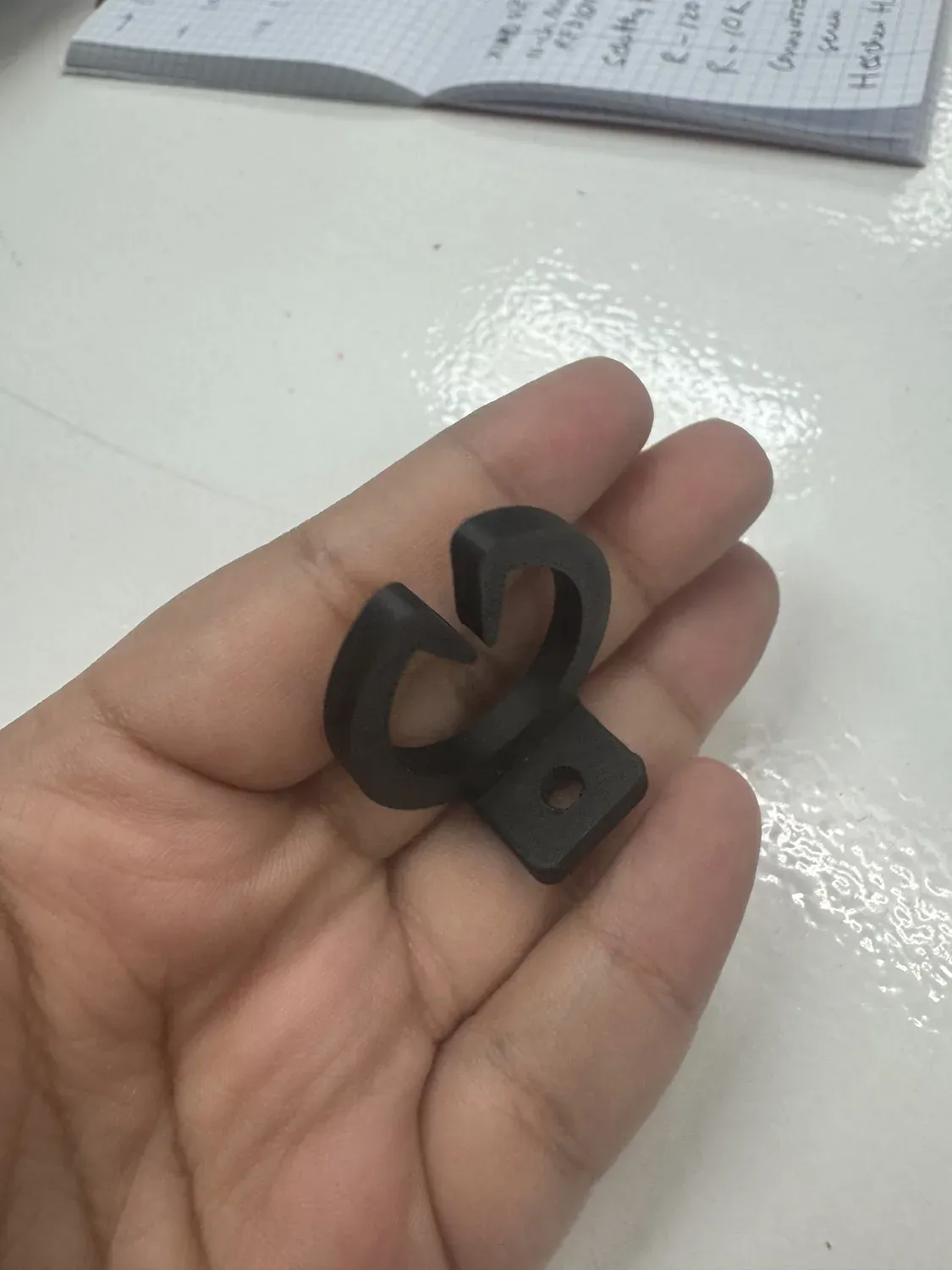 3D-printed black cable organiser clip with a mounting tab and screw hole, held in hand