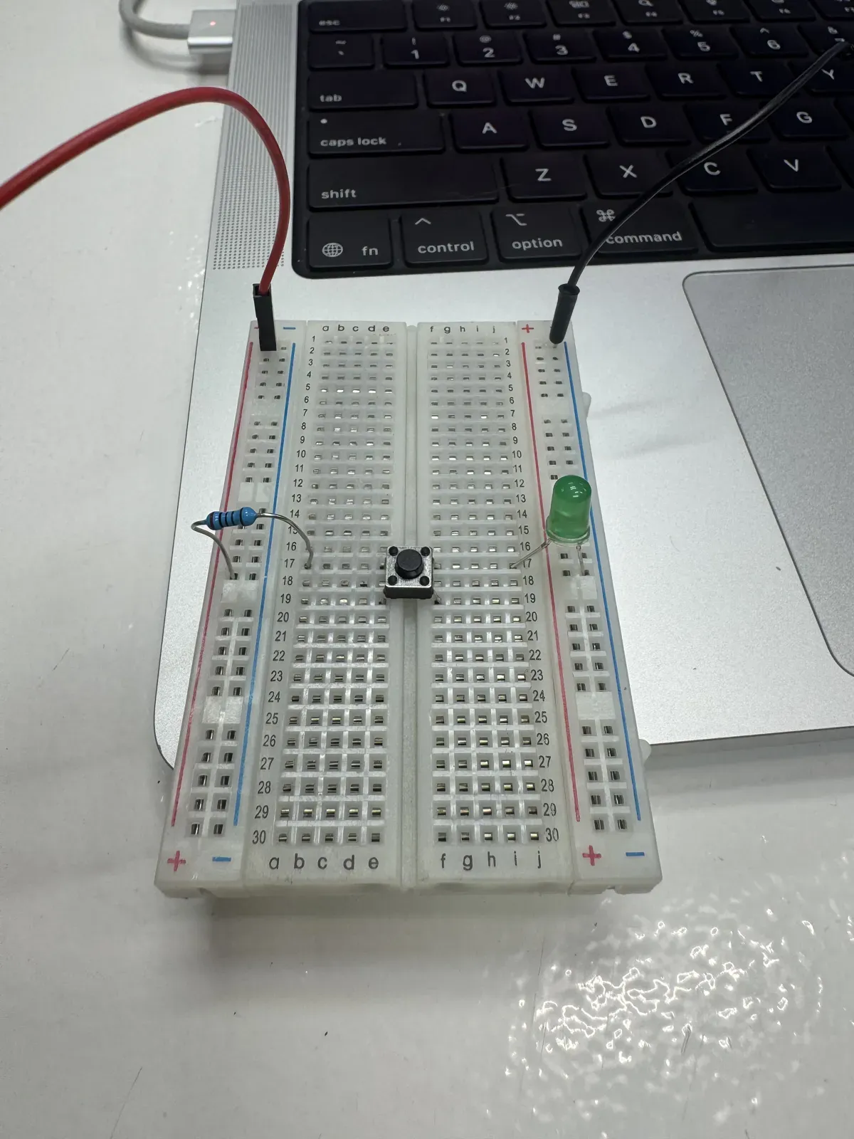 Assembled breadboard circuit with resistor, button, and green LED connected to laptop USB power