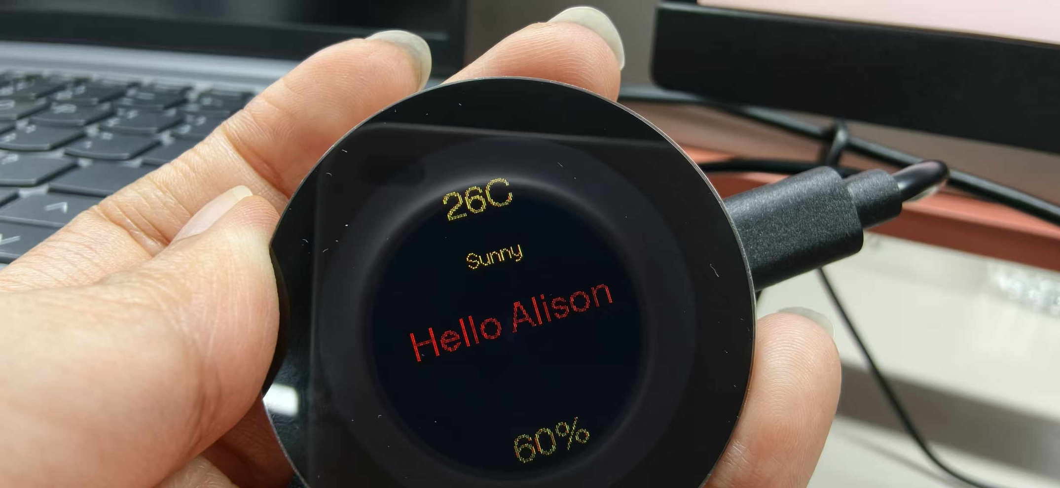 Round display showing “Hello Alison” with temperature, weather line, and humidity