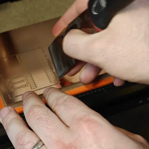 Using a scraper to pry the finished PCB off the double-sided tape on the machine bed
