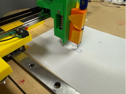 Title: image21.jpeg - Description: Pen carriage of the Coloured Plotter putting down its first deliberate line — the green PLA pen-holder bracket, sprung Z-lift, and orange masking tape used to dial in pen height.