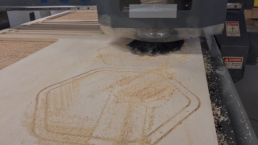 The CNC router cutting the rounded-hexagon ring profiles into a plywood sheet, dust shoe down and sawdust around the cut.