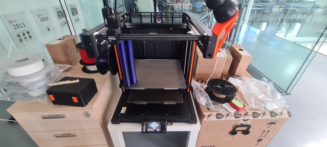 An Original Prusa XL printing a tall blue PETG body, filament spools and tools around it in the fab lab.
