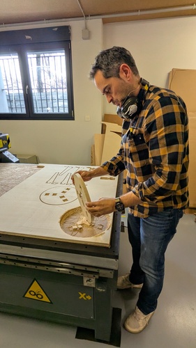 Lifting one of the freshly cut stool pieces from the CNC table, with sawdust visible around the cut perimeter.