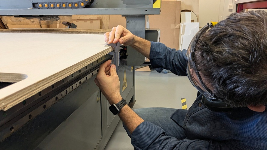 Measuring the edge of the plywood board with a digital caliper against the machine rail, wearing safety glasses with ear protection nearby.