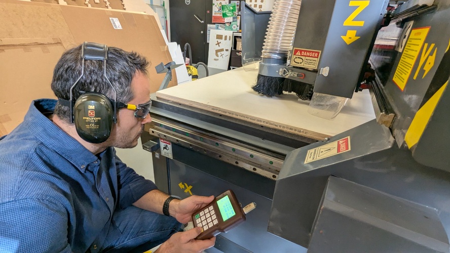 Wearing full PPE — ear protection and safety glasses — verifying the job has started correctly on the RichAuto A11, with the USB loaded, the spindle access cover closed, and one finger near the cancel button.