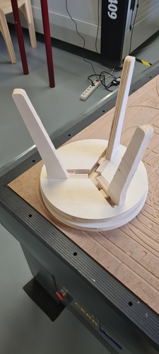 The assembled stool standing on the CNC table with the two plywood legs crossing and the circular seat base visible.