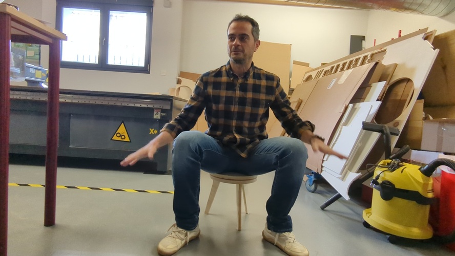 Person sitting on the flat-pack plywood stool in the Fab Lab León CNC room, arms open, stool stable under full weight.
