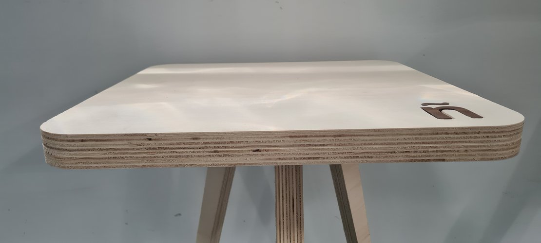 Low-angle close-up of the tea table top edge showing the plywood layers, the rounded corner profile, the ñ inlay in darker wood, and the three leg tenons visible underneath.