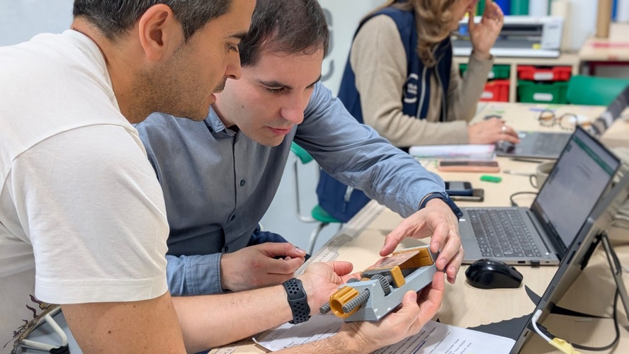 Adrián and the student examining the partially-soldered board held in the 3D-printed PCB vise, a laptop and notes on the bench.