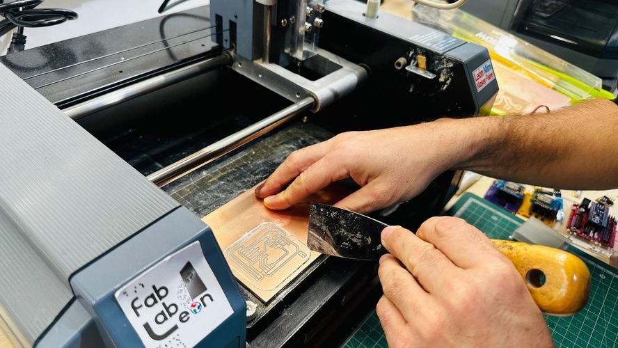 Hand sliding a flat spatula under one edge of the milled board on the MDX-20 bed, Fab Lab León logo on the machine.