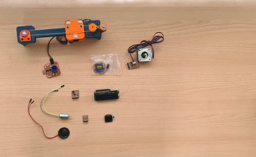 Collection of output devices laid out on a wooden table: speaker, DC motor, small custom PCBs, OLED display, LCD display, servo motor in packaging, and a NEMA 17 stepper motor with A4953 driver board. An orange and blue robotic arm assembly is visible on the right.