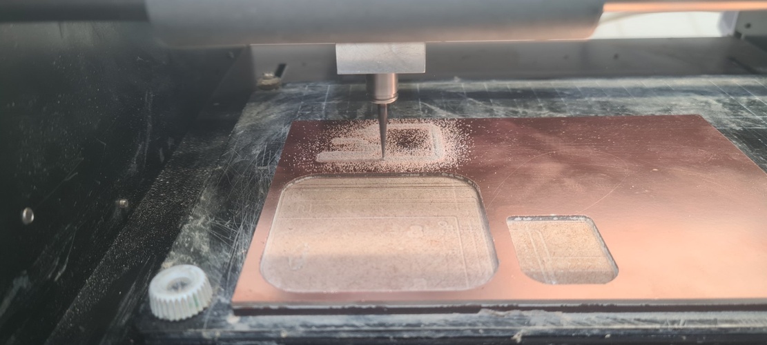 Roland MDX-20 milling machine mid-job on a copper FR1 board. The endmill is cutting traces around an IC pad area, with fine copper dust visible. A second smaller board outline is visible to the right on the same copper sheet.