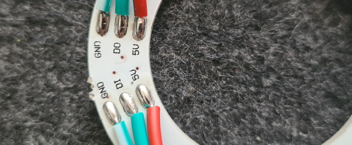 Close-up of the WS2812B ring showing the silkscreen labels GND, DI, 5V on the input side and GND, DO, 5V on the output side, with three coloured wires soldered to the input pads.