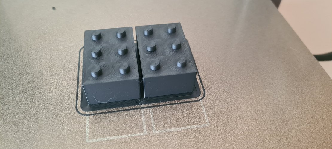 Two black PLA prints of the 2x3 brick on the textured Bambu A1 build plate, fresh out of the printer. The prints share a brim around the base. The visible top surface shows the printer's layer texture on both the brick body and the studs.