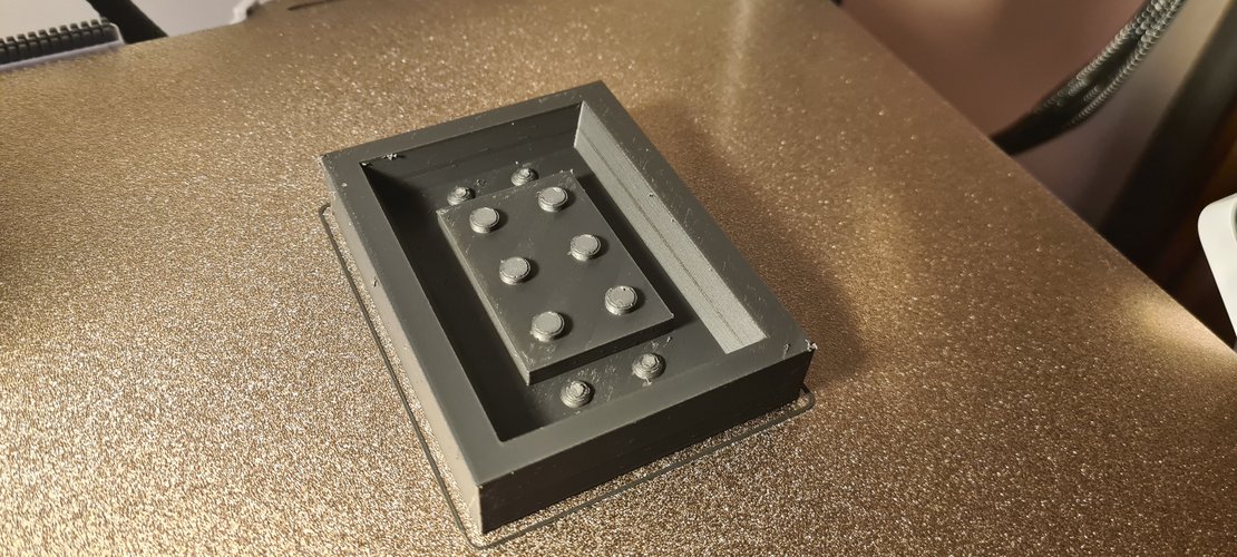 Top-down photo of the FDM-printed PLA master in grey, sitting inside its integrated casting box. The brick's six studs are clearly visible on top. Two small witness studs sit in one corner of the box floor, near the master. The print rests on a metallic textured desk surface.