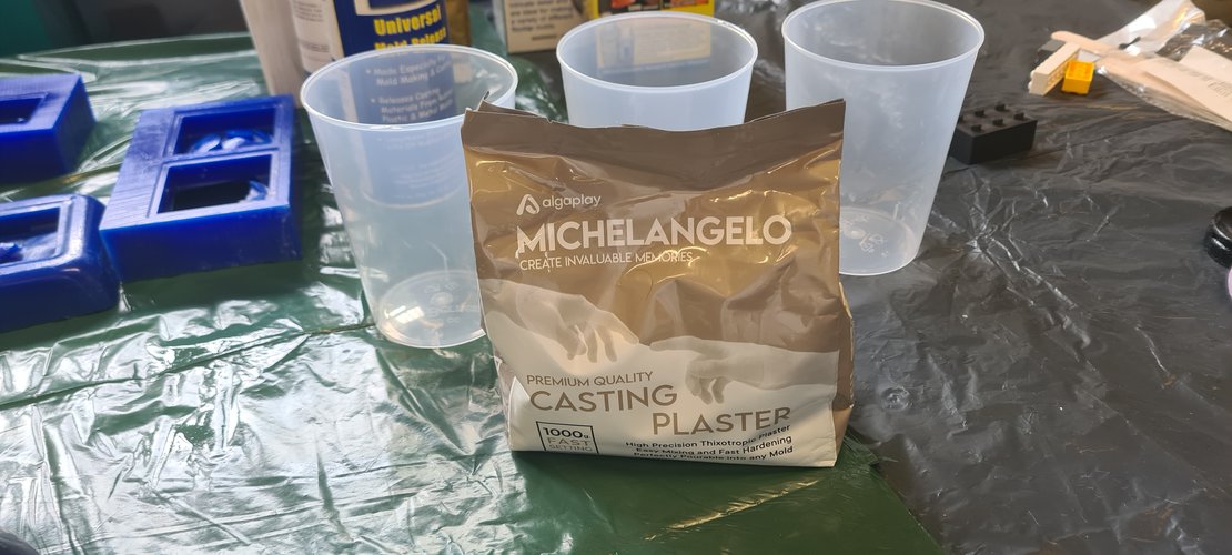 The Algaplay Michelangelo plaster bag set up between three measuring cups on the workbench. A blue 3D-printed mould sits to the left for reference. The Smooth-On Universal Mold Release can is in the background.