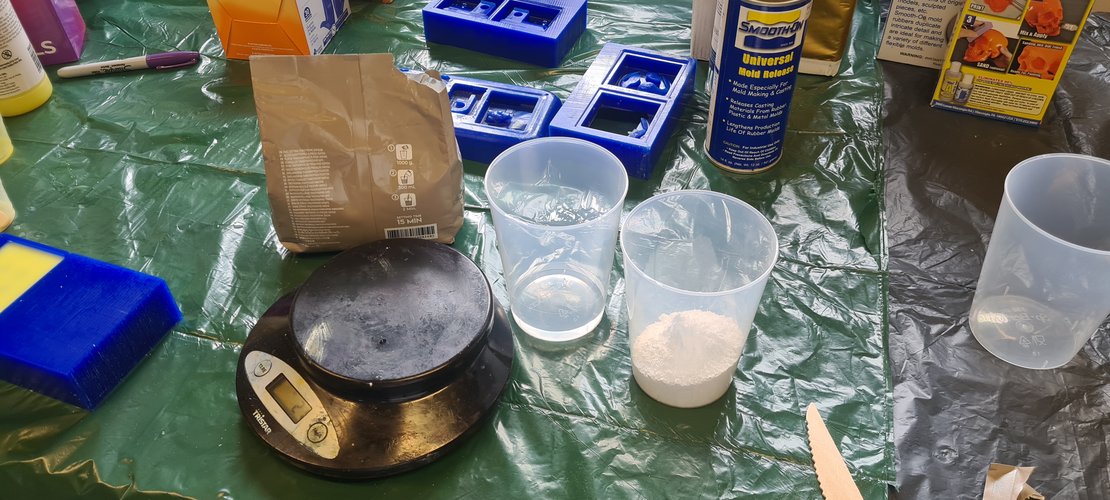 The plaster bag with the back side visible showing the printed instructions: 1000 g, 300 ml, 2 min mixing. Two cups in front of the bag — one with clear water, one with white plaster powder. The kitchen scale sits to the left.