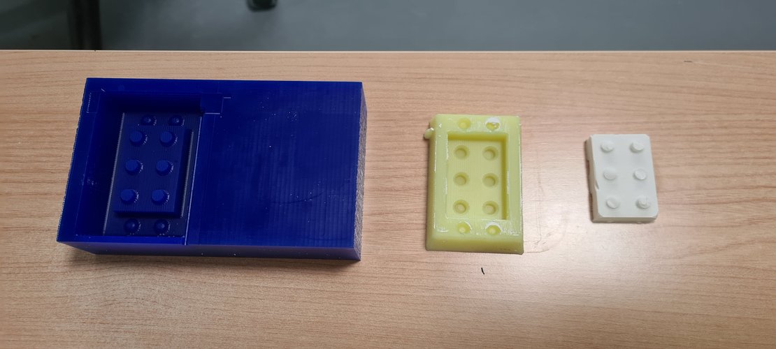 Side view with the three artefacts of the full process aligned on a wooden desk. Left: the blue wax block with the wax master inside it. Centre: the yellow silicone mould, demoulded. Right: the white plaster cast of the brick, free-standing, showing six studs of varying definition.