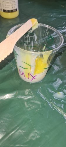 Compostable cup labelled 'MIX' with combined silicone, partly streaky from incomplete mixing. A wooden stir stick rests on the rim with silicone running down it. The cup sits on a green plastic sheet protecting the workbench.