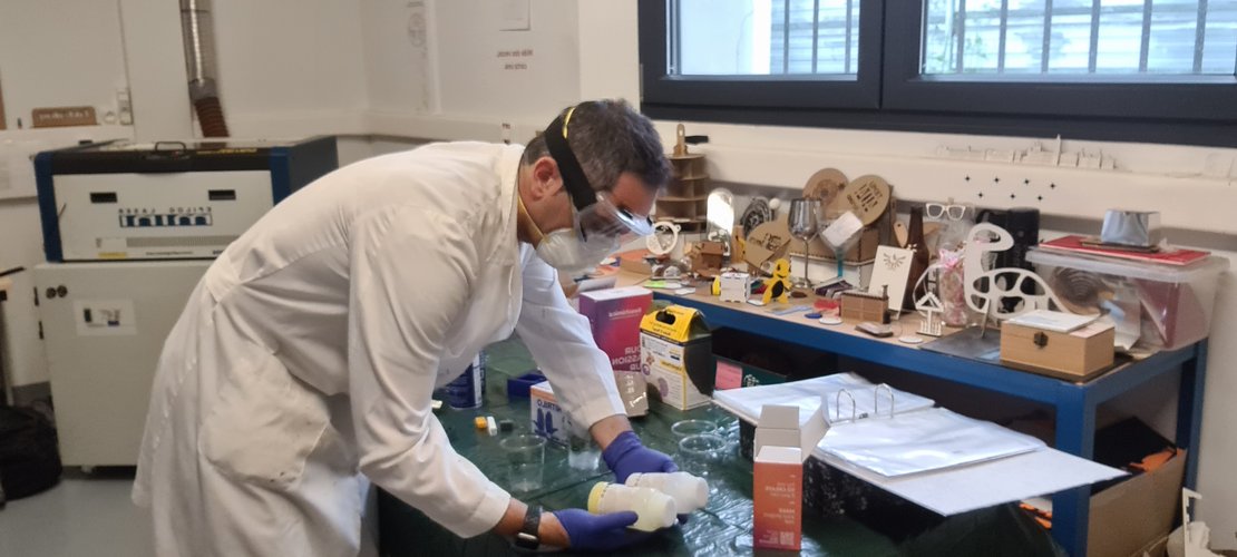 Photo of me at the Fab Lab León workbench wearing PPE — lab coat, safety goggles, FFP2 mask, nitrile gloves — handling the two silicone bottles.