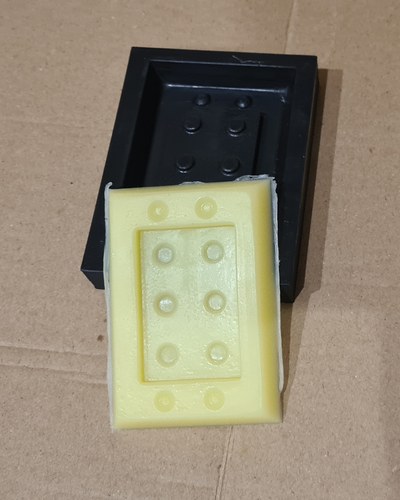 The demoulded yellow RPRO 30 silicone mould resting next to the black PLA master and its casting box, on cardboard. The brick and stud detail transferred cleanly into the silicone.