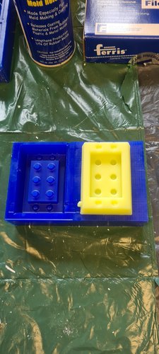 Two elements after demoulding, on a green plastic sheet. Left: the blue wax block with the wax master sitting back inside its original cavity, the six blue studs clearly visible. Right: the cured yellow silicone mould, free-standing, showing a rectangular cavity with six cylindrical hollows in the floor where the master's studs had been.