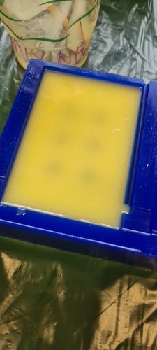 Top-down view of the blue wax block holding the master, with yellow silicone freshly poured into the cavity. The silicone is still liquid and partly translucent — the wax studs are visible underneath as faint shadows. The MIX cup with the leftover mixed silicone sits at the top of the frame.