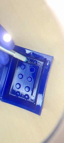 Close-up of the wax master under an LED work lamp, with a precision cutter blade trimming wax material around one of the corner studs. The release agent gives the surface a wet sheen.