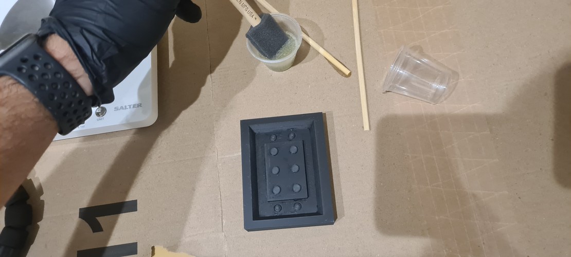 A foam brush loaded with mixed XTC-3D resting over the cup, next to the PLA master and its integrated casting box on the cardboard surface, just before brushing the coating on.