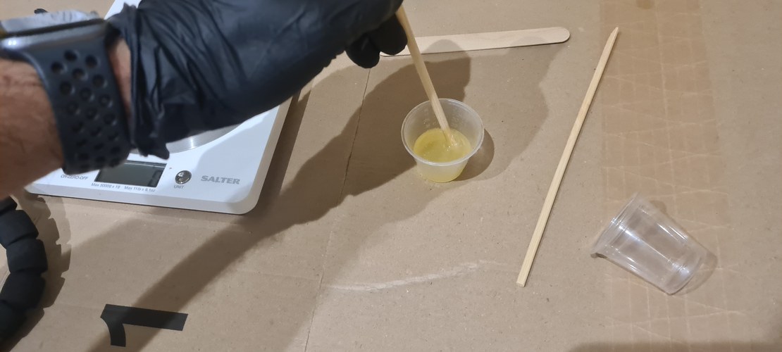 Gloved hand stirring the mixed XTC-3D, a pale yellow liquid, in a small plastic cup with a wooden stick. The cup sits on a Salter kitchen scale used to weigh parts A and B. A foam brush and wooden skewers lie on the cardboard work surface.