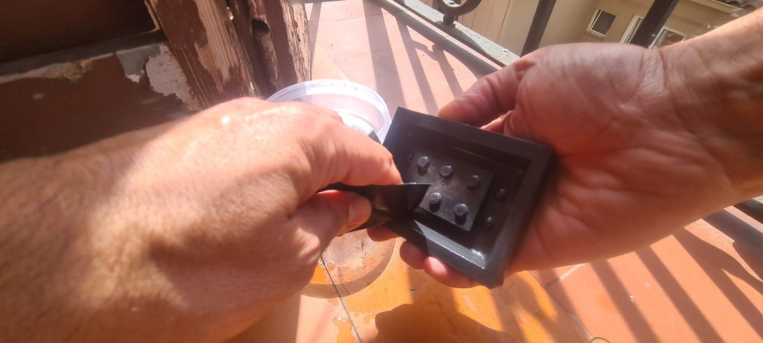 Two hands outdoors on a tiled balcony: one holds the coated PLA master, the other a folded sheet of sandpaper, with a cup of water nearby for wet-sanding.