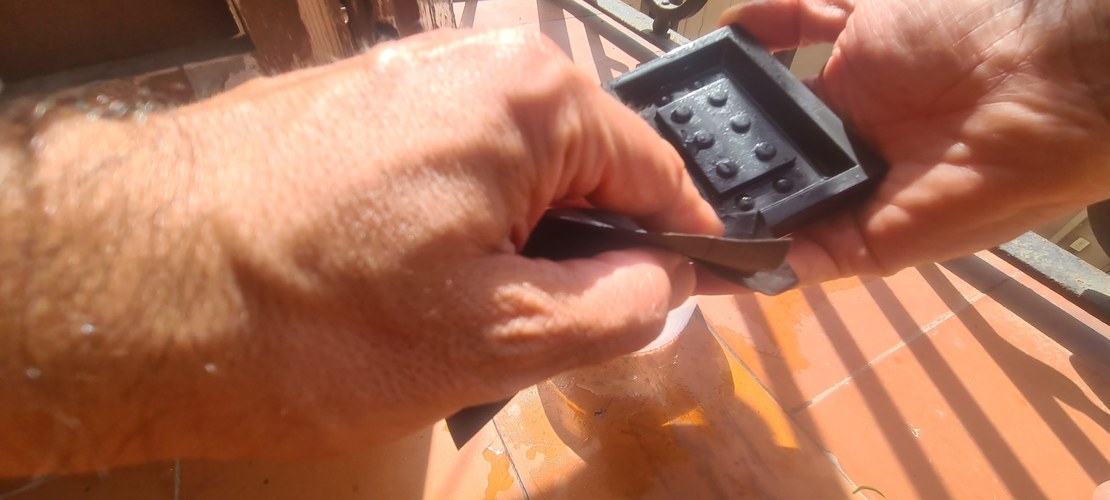 Close-up of both hands wet-sanding the coated PLA master, running a folded sheet of sandpaper across the studded face.