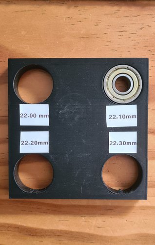 A 3D-printed black PLA test piece with four holes at 22.00, 22.10, 22.20 and 22.30 mm, with a 608ZZ bearing sitting on top of the 22.10 mm hole showing the chosen fit.