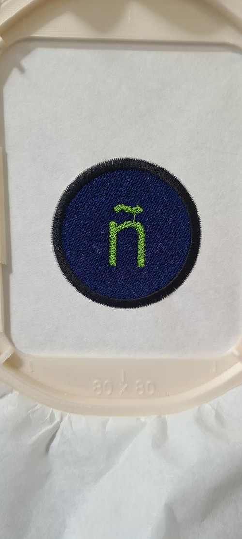 The finished embroidered patch in natural light, still in the 80x80 hoop: blue fill, lime-green ñ, dark satin border.