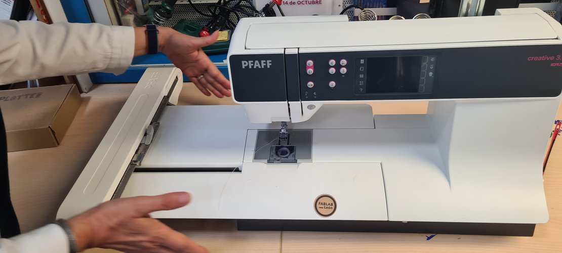Nuria pointing at the PFAFF machine with the embroidery arm attached, FABLAB León sticker visible.