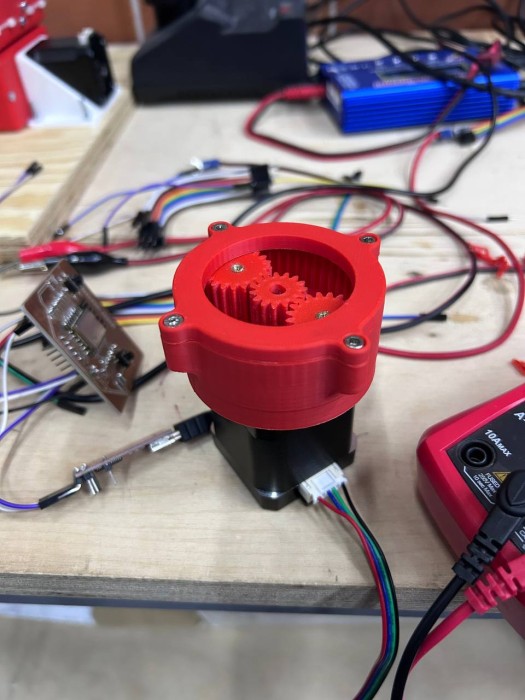 3D printed reducer parts assembled on Nema 17 motor — red PLA housing with planet gears visible through open top, three gears meshing correctly, motor cable visible, lab workbench in background with electronics