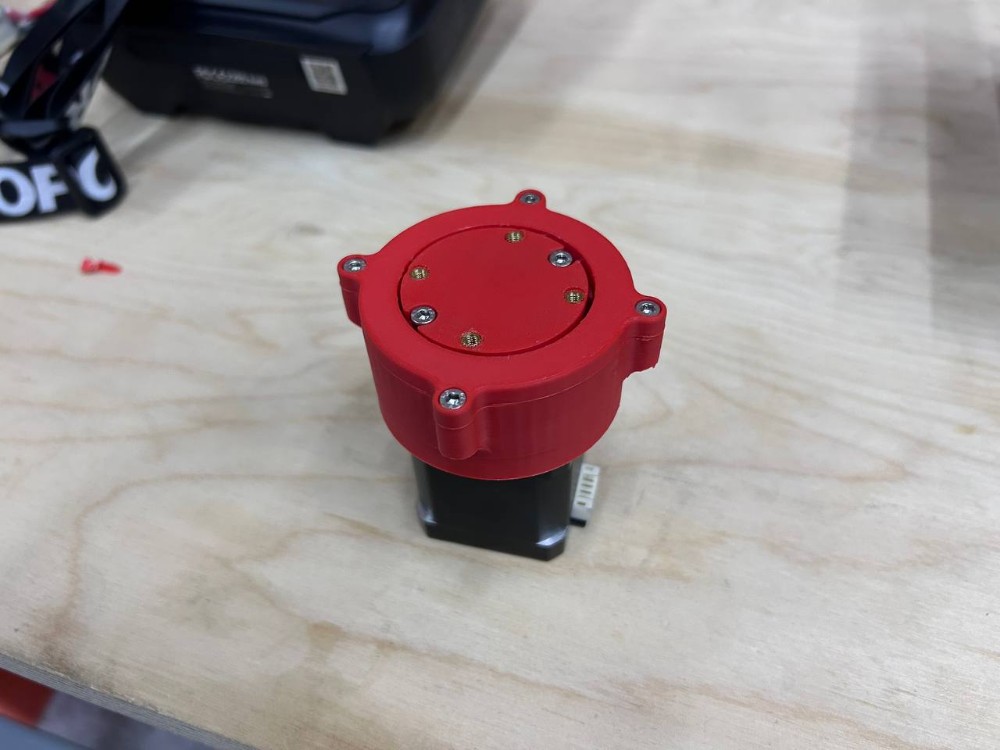 Completed reducer assembly — red PLA housing with top cover installed, six M3 bolts with brass inserts visible on top face, four side mounting ear screws, Nema 17 motor at base on wooden workbench