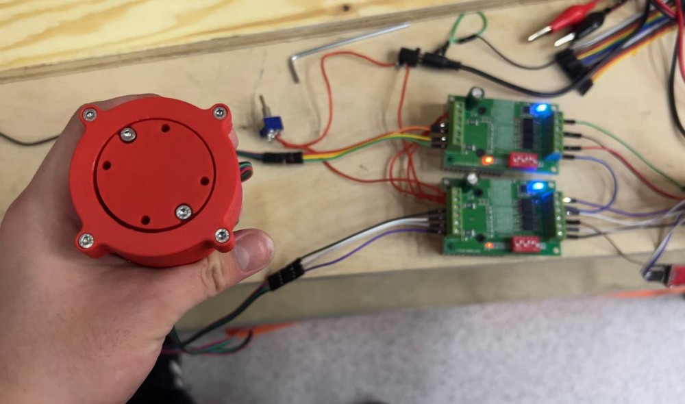 Completed reducer held in hand — top view showing output face with six M3 bolts, connected to TB6600 stepper motor driver boards on wooden panel in background, blue LED indicators visible on drivers