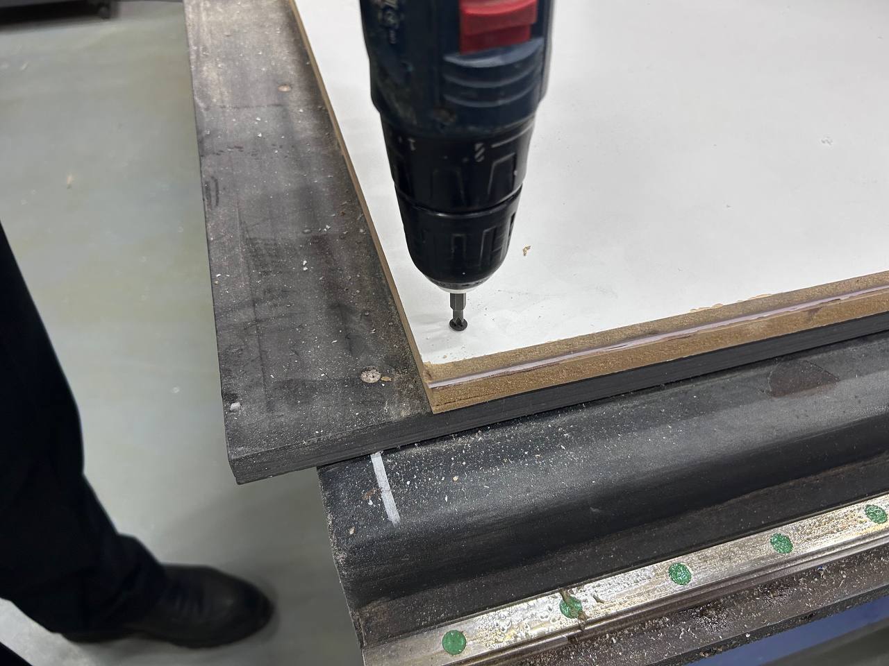 CNC machine bed — screwdriver drilling a self-tapping screw through the corner of the white MDF sheet into the sacrificial bed, machine rail and green alignment dots visible