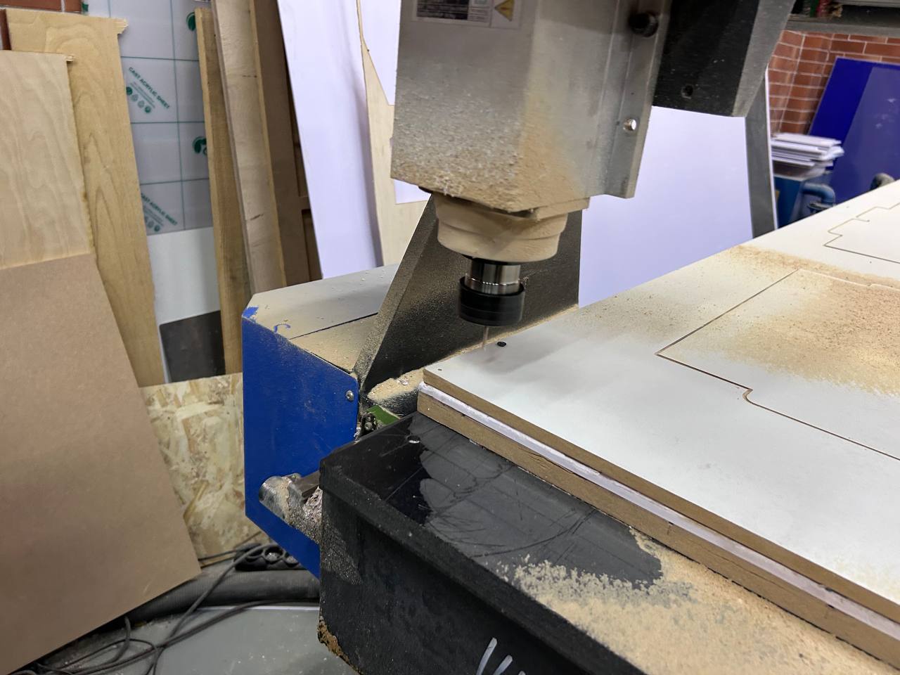 CNC router spindle close-up — router head lowered onto the white MDF sheet, bit visible cutting into material, dust accumulation visible on the board surface, plywood pieces leaning against wall in background