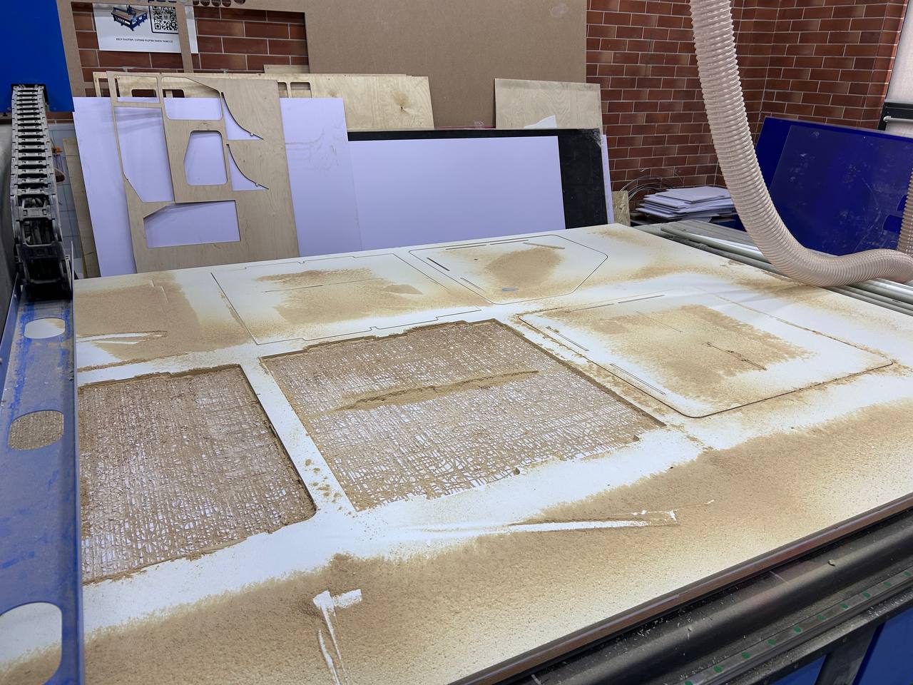 CNC machine bed after completing all cuts — white MDF sheet with all panel cutouts complete, sawdust covering the surface, cut pieces visible in their slots, vacuum hose visible, previous MDF cuts leaning against wall