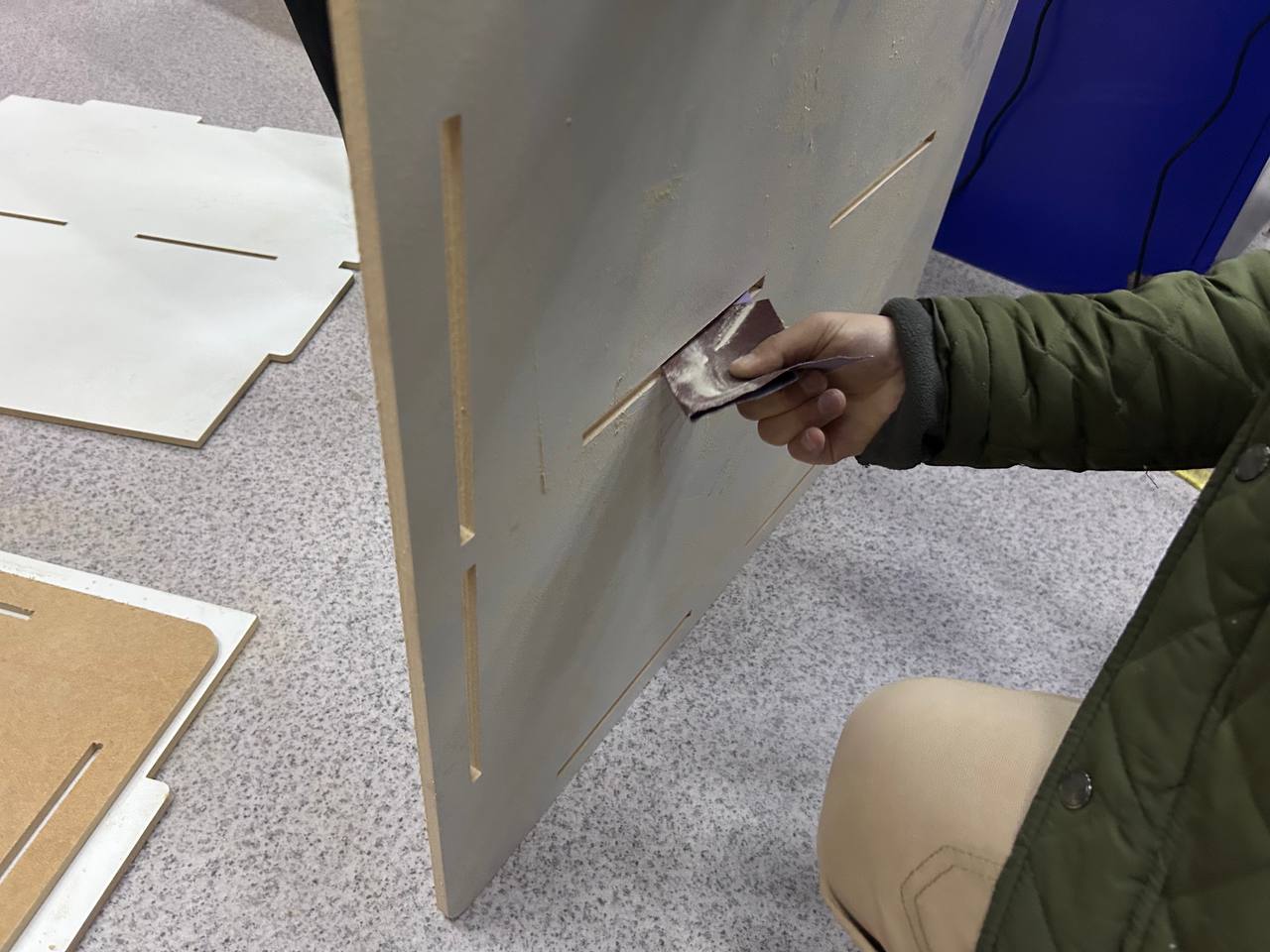 Sanding the milled MDF piece — a hand holding sandpaper is sanding the edge of a large vertical MDF panel, slot cutouts clearly visible, other panels visible on the floor to the left